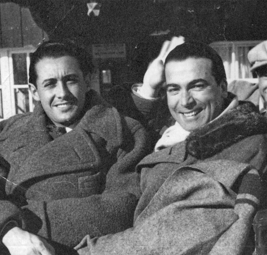 Cristóbal Balenciaga – Maestrul Tăcerii, Arhitectul Formei și Misterul Spaniol 3 Fotografie alb-negru, realizată în aer liber, care îl înfățișează pe Cristóbal Balenciaga, celebrul designer de modă spaniol, zâmbind cald alături de Wladzio d’Attainville, partenerul său de viață și om de încredere.